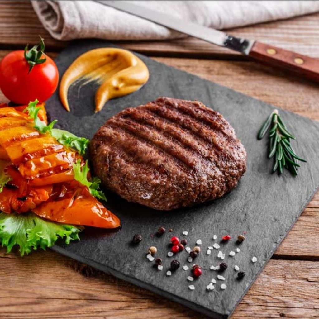 A grilled beef patty served on a slate platter with roasted peppers, fresh lettuce, a tomato, and mustard.
