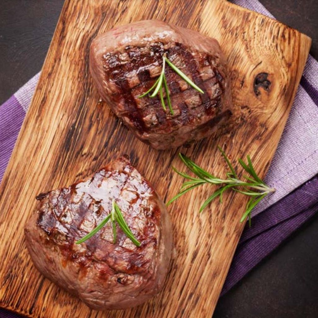 Two seared beef steaks garnished with rosemary sprigs on a wooden cutting board over a purple cloth.