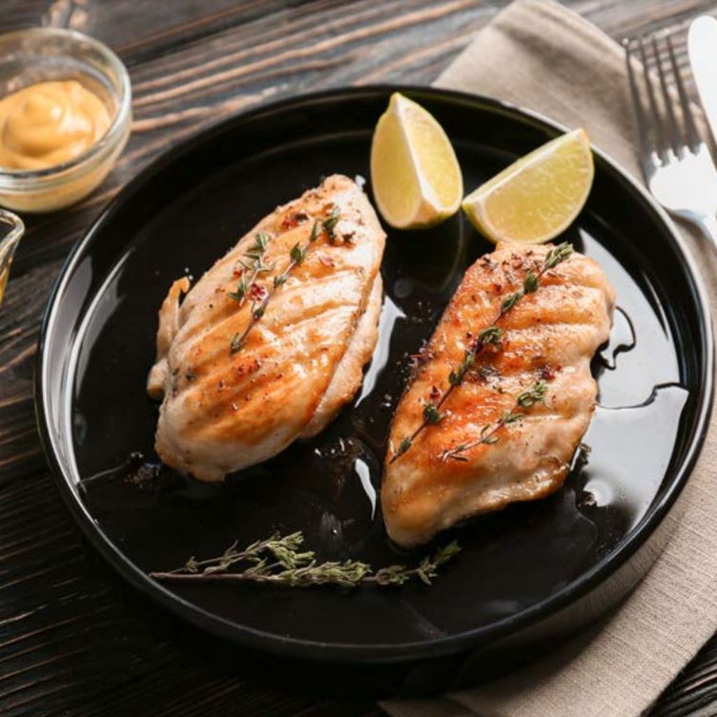 Two grilled chicken breasts garnished with thyme sprigs and lime wedges on a black plate.