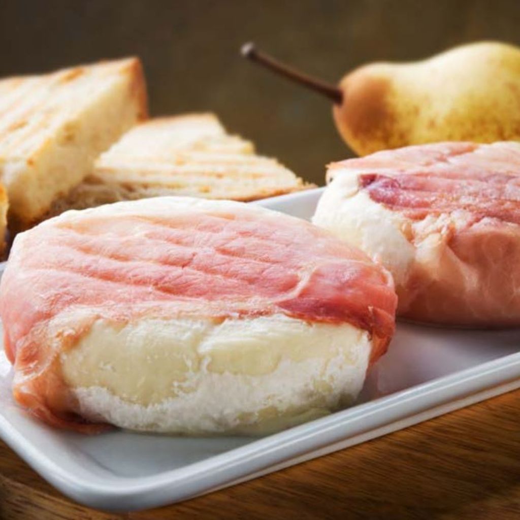 Two rounds of grilled Tomino cheese wrapped in speck, served on a white plate with toasted bread and a pear.