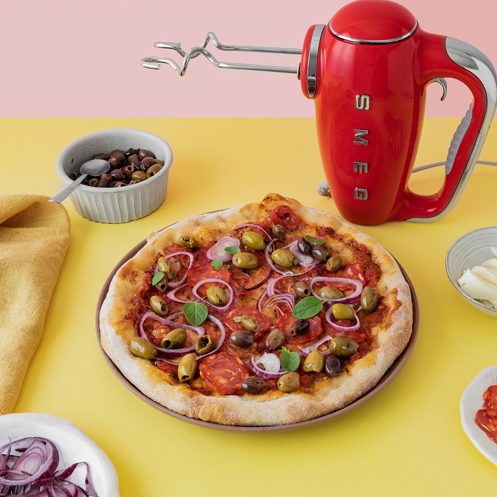 Red Smeg hand mixer next to a fresh pizza topped with red onion, green and black olives, and spicy salami.