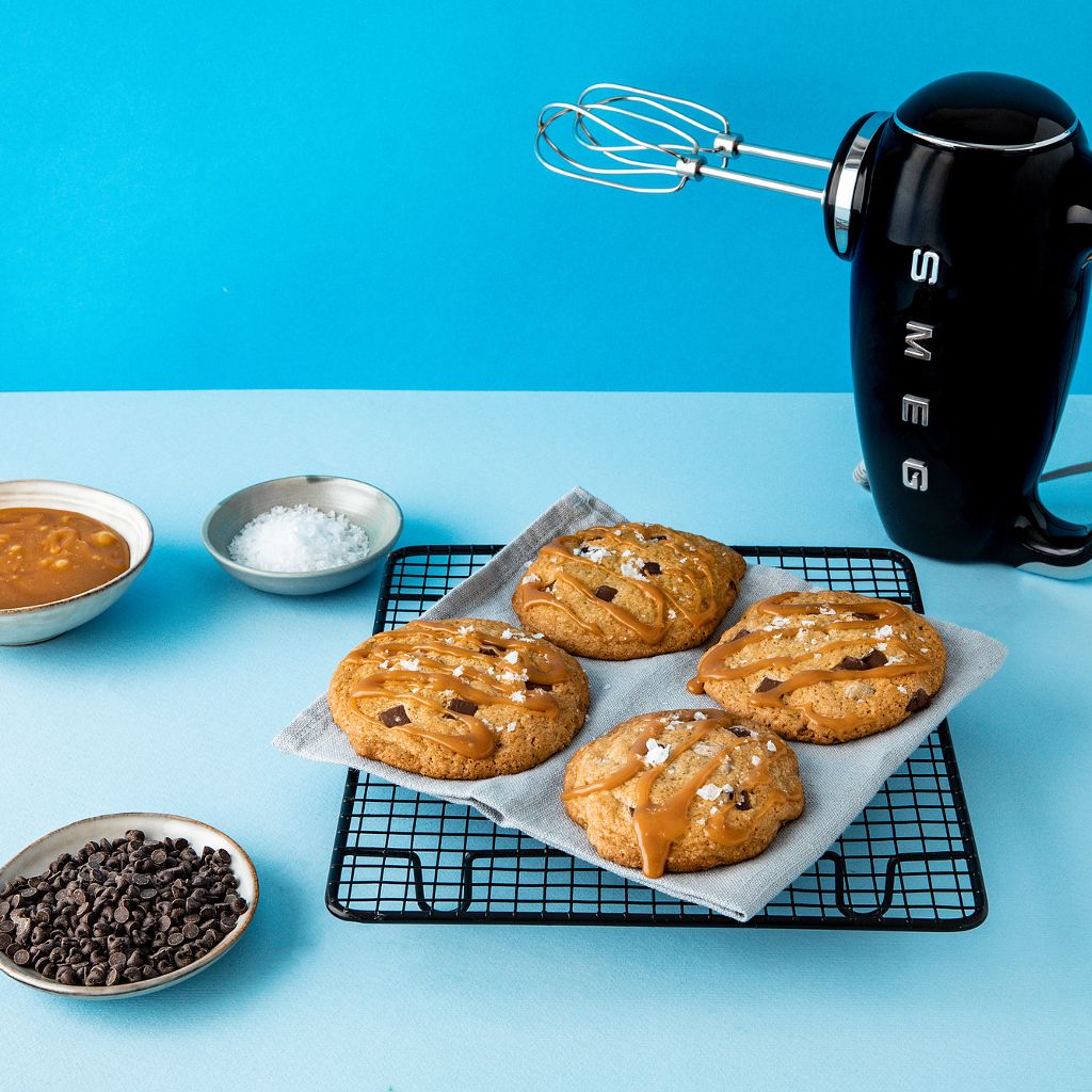 Four cookies with caramel drizzle and sea salt on a wire rack next to a Smeg hand mixer and bowls of salt.