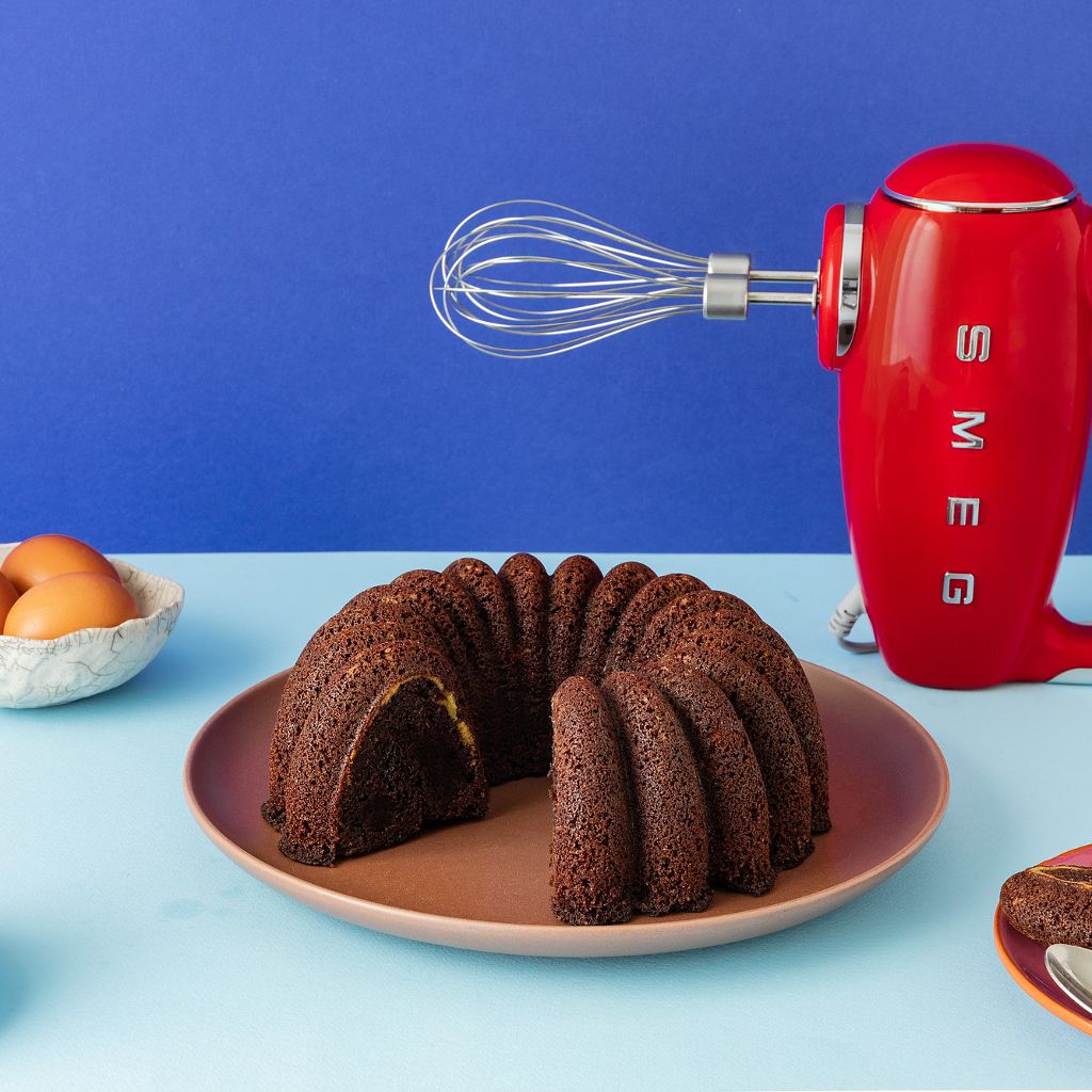 A red Smeg hand mixer with whisk attachment stands beside a sliced chocolate bundt cake on a blue background.
