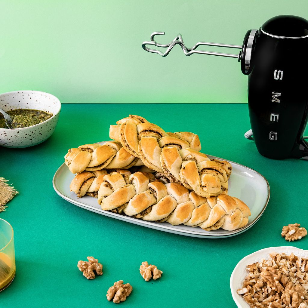 A black Smeg kettle sits behind a platter of golden pastry braids filled with pesto and walnuts on a green surface.