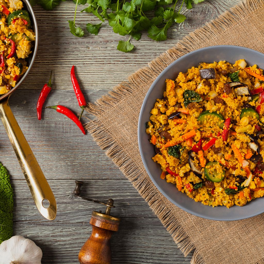 A gray bowl of golden saffron couscous mixed with zucchini, peppers, and carrots, served with fresh red chilies.