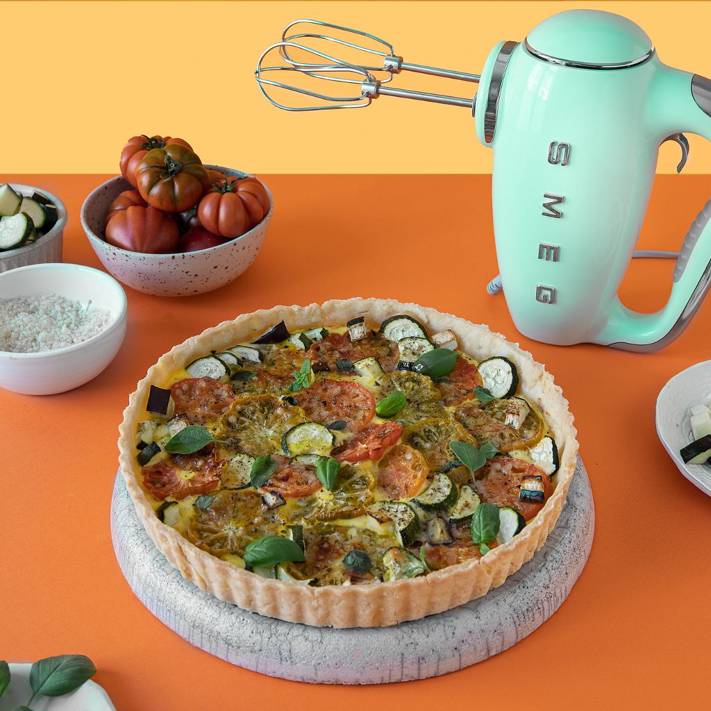 A mint green Smeg hand mixer stands behind a savory tart filled with sliced tomatoes and zucchini on an orange background.