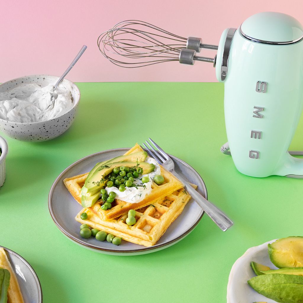 Savoury waffles topped with cream cheese and peas on green backgrounds, next to a pastel green Smeg hand mixer.