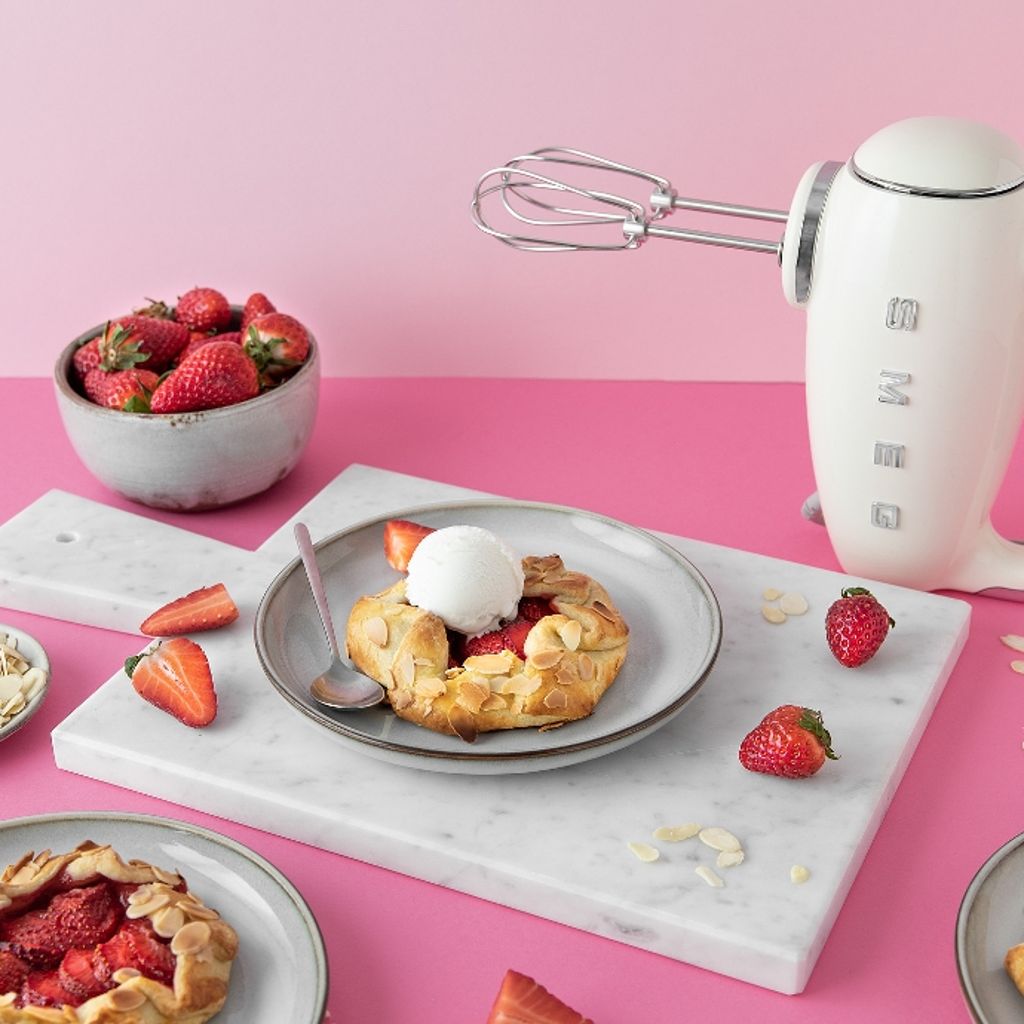Three strawberry mini galettes on grey plates, one topped with a scoop of ice cream, next to a white Smeg kettle on a pink background.