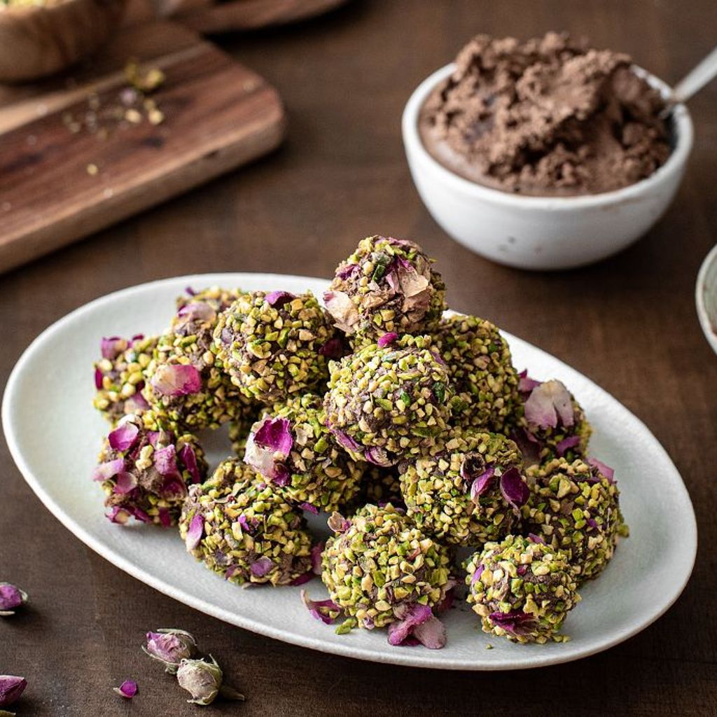 Decadent chocolate truffles coated in crushed green pistachios and dried pink rose petals on a white oval platter.