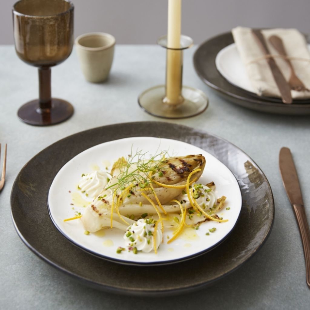 Plated grilled endive with dollops of white cheese, orange zest, and pistachios on a table with candles and glassware.