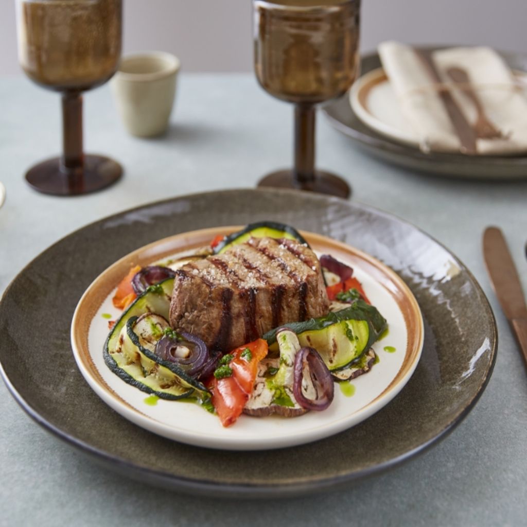 Grilled beef fillet over zucchini, onions, and peppers on a white plate, set on a table with candles and wine glasses.