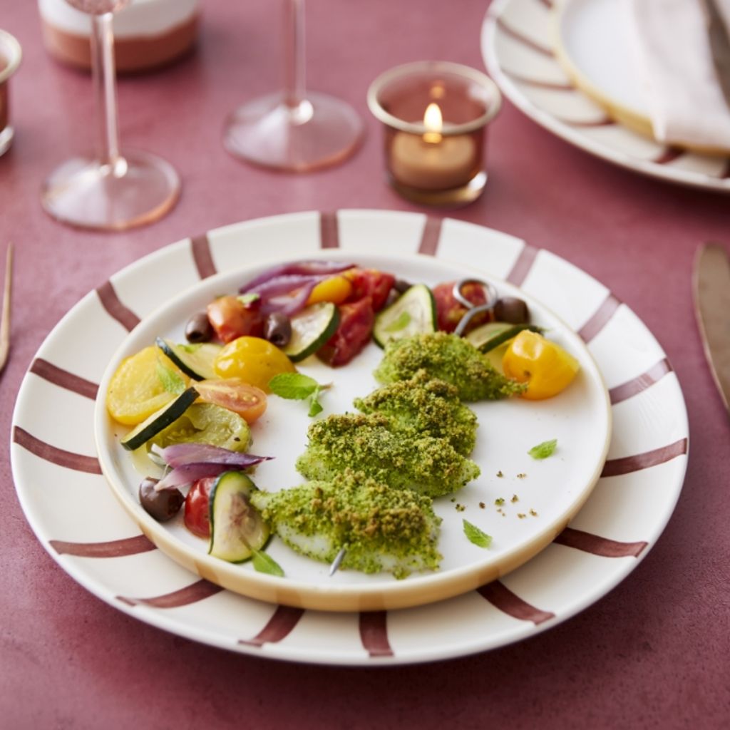 Skewer of green-crusted squid with tomatoes, olives, and zucchini on a striped plate with candles.