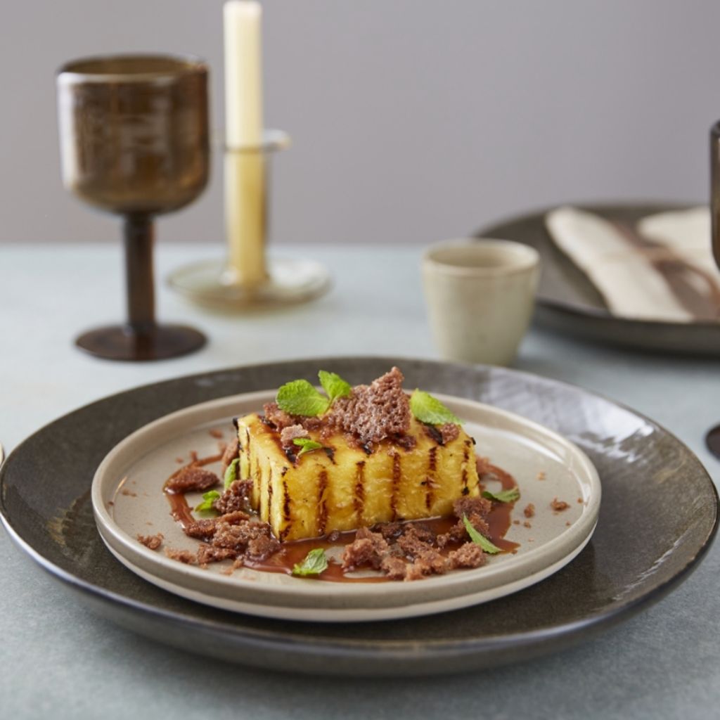 A golden-brown grilled pineapple slice topped with buttery crumble, caramel sauce, and fresh mint on a white plate.