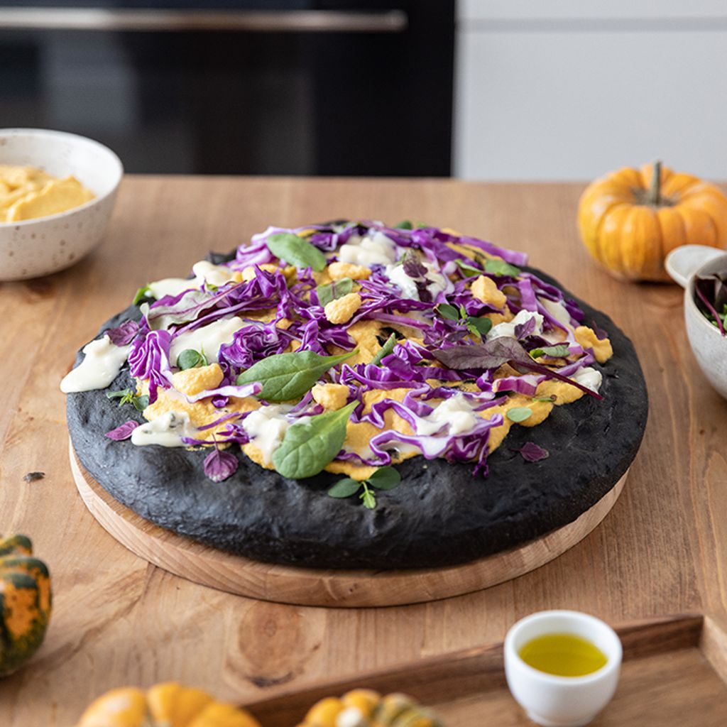 A round, jet-black focaccia on a wooden board topped with orange pumpkin hummus, purple cabbage, spinach, and white gorgonzola.