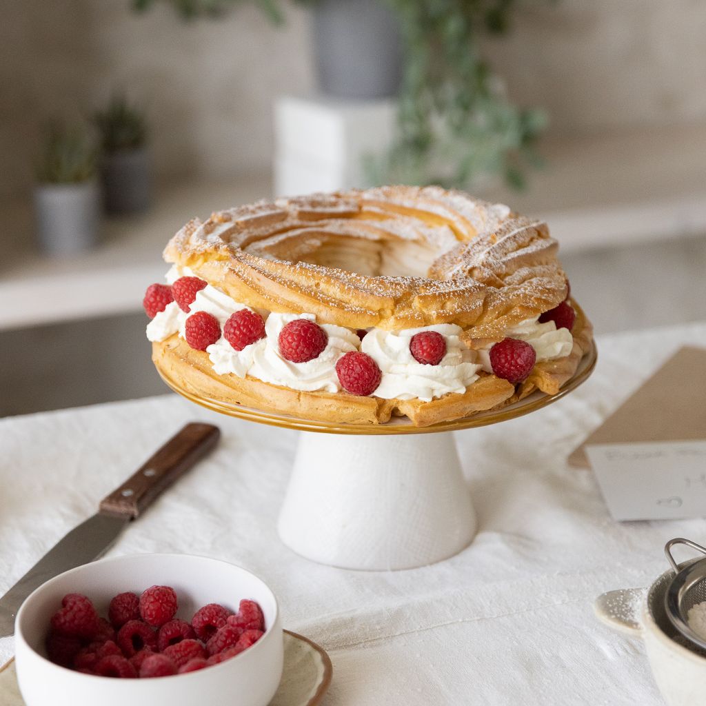 A Paris-Brest cake on a cake stand, filled with swirls of white cream and fresh raspberries, on a beautifully laid table.