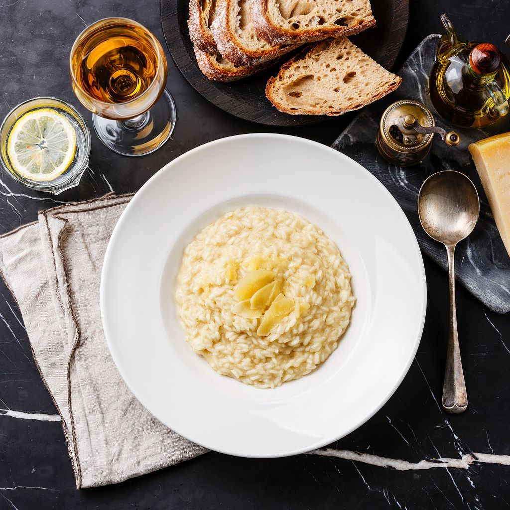 Creamy risotto with melted butter and parmesan served in a white plate with bread, olive oil and wine on a dark marble table