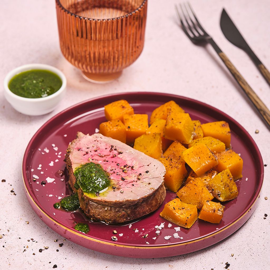 Plated sliced roast pork with green herb sauce and roasted pumpkin cubes on a ceramic dish
