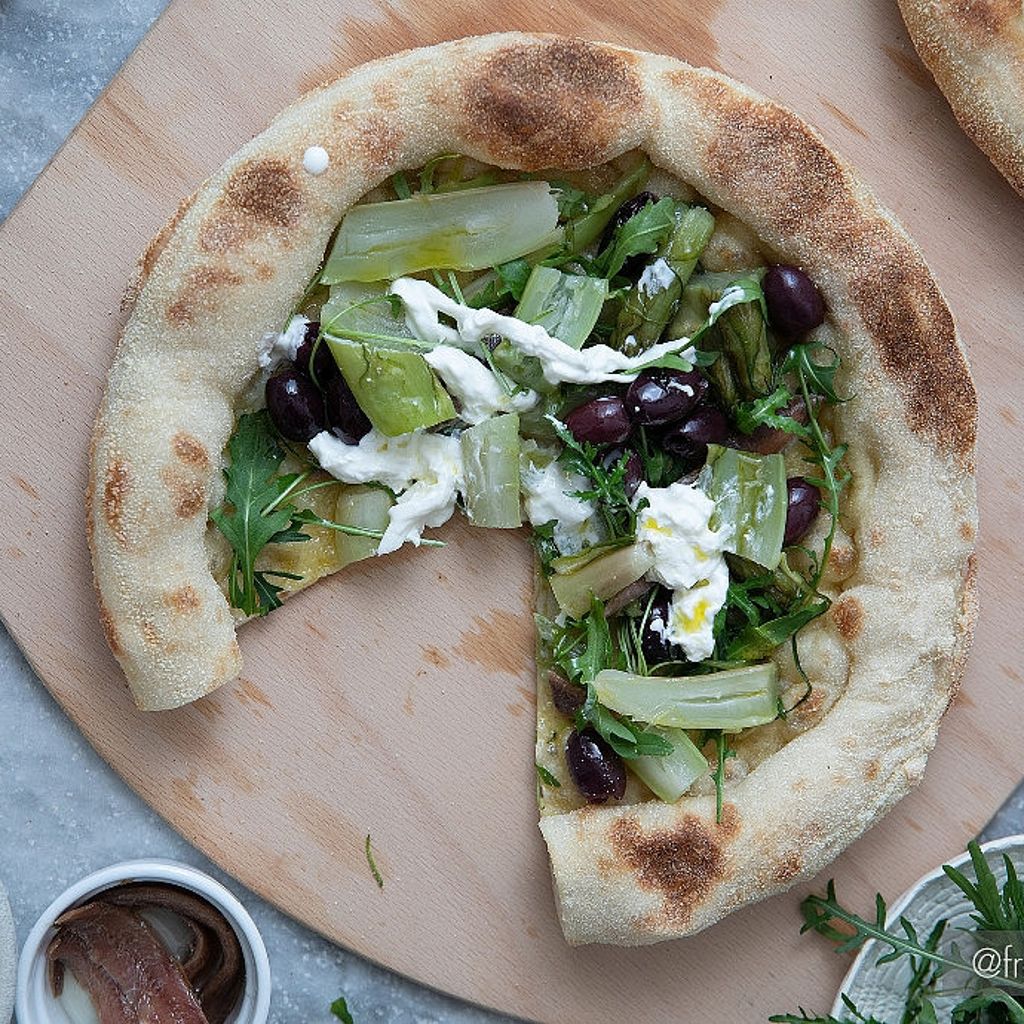 A pizza topped with chicory, black olives, and fresh burrata cheese on a wooden board, with side ingredients visible.