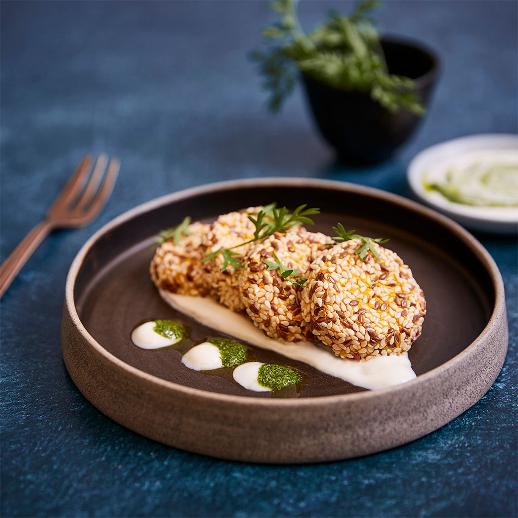Three round falafels topped with sesame seeds, served on a bed of white cream with a drizzle of green sauce, on a dark grey plate.