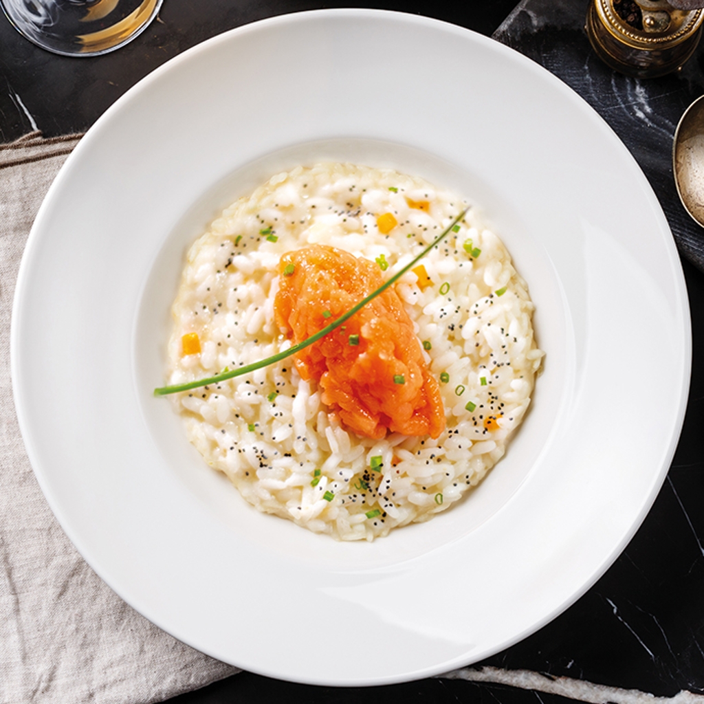 White risotto with a salmon tartare centre, chives and poppy seeds, served on a white plate set against a black marble background.