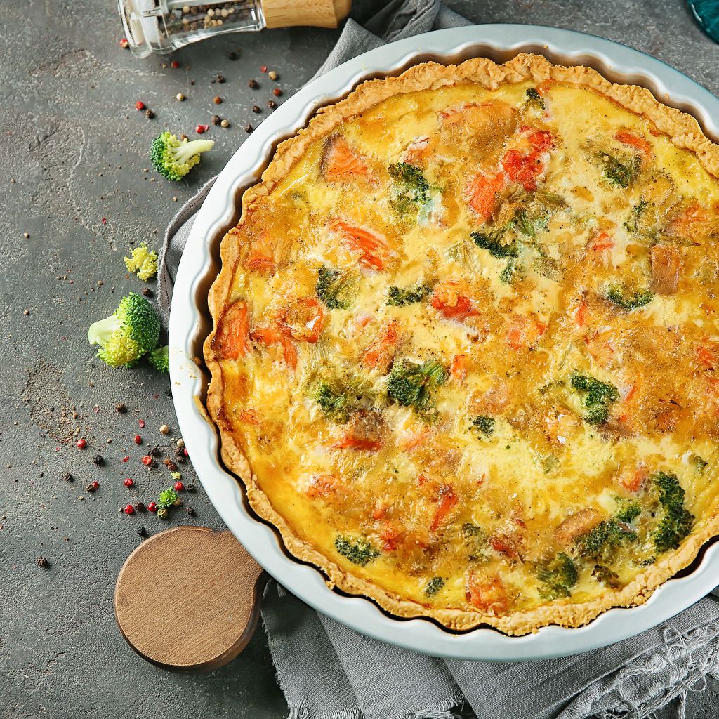 Top-down view of a golden-brown quiche with visible vegetables in a white ceramic dish on a grey stone surface.