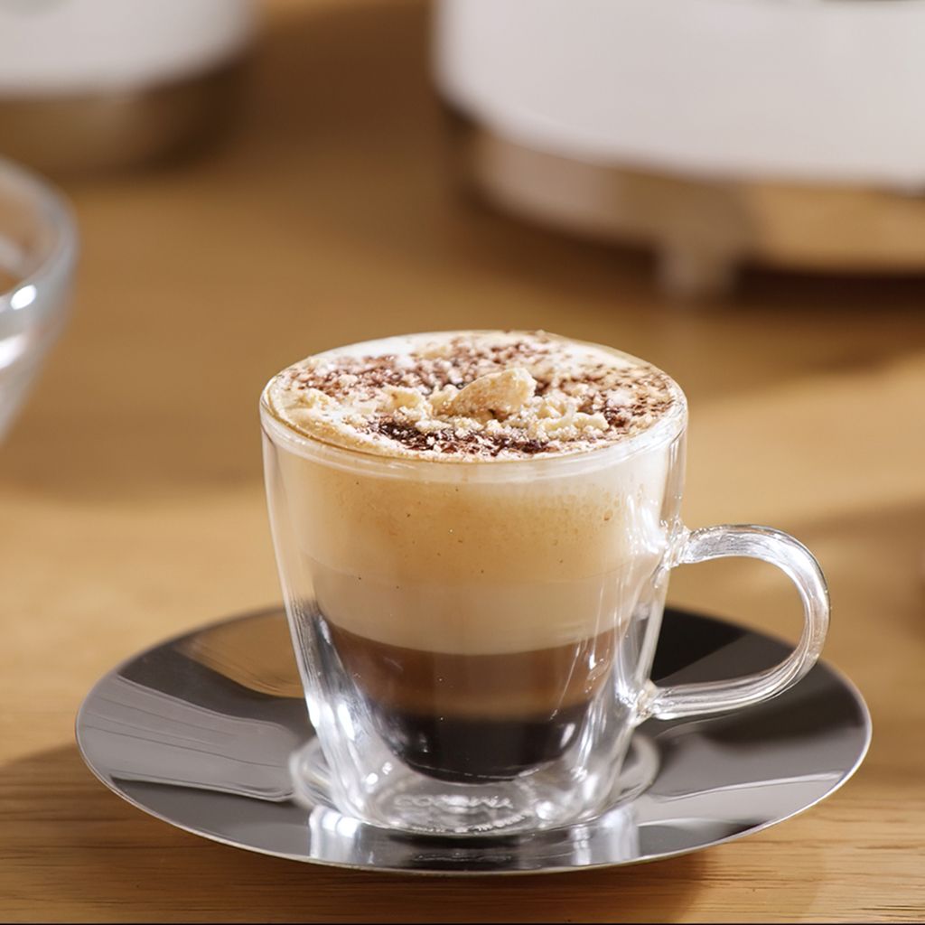 A layered choco macchiato served in a clear glass cup on a reflective saucer, topped with creamy foam and chocolate crumbles