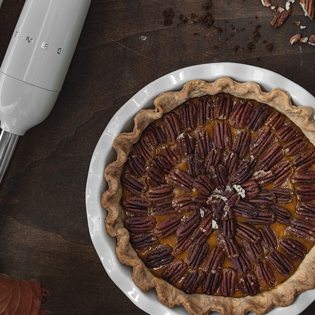 A pumpkin tart topped with pecans in a white baking dish, next to a white Smeg hand blender.