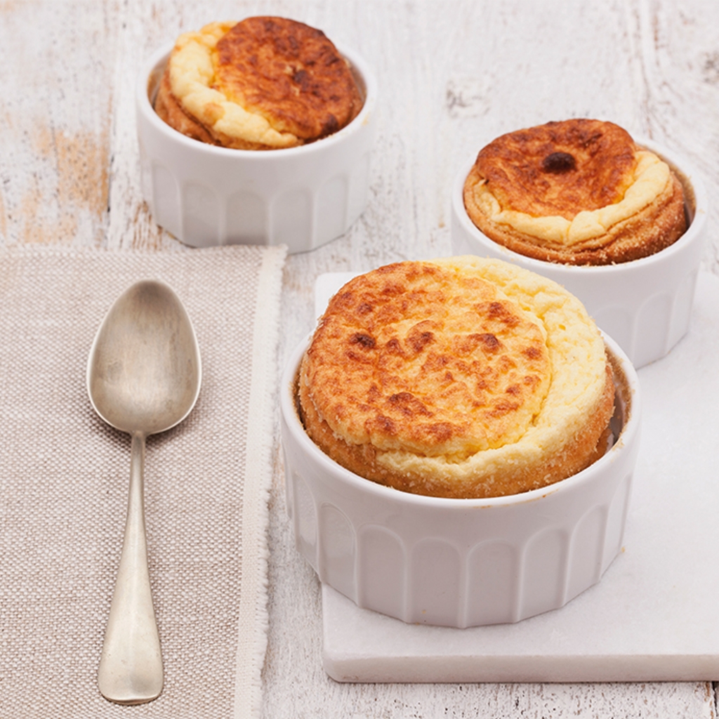 Three golden soufflés in white paper cases on a light-coloured wooden board, accompanied by a silver spoon.