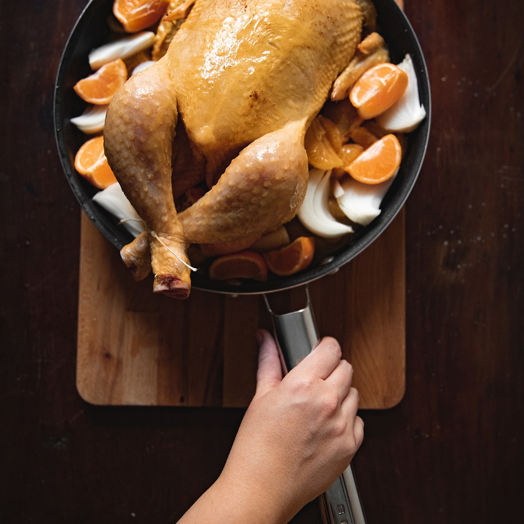 A whole capon in a roasting tin, surrounded by clementine segments and white onion, on a wooden chopping board.