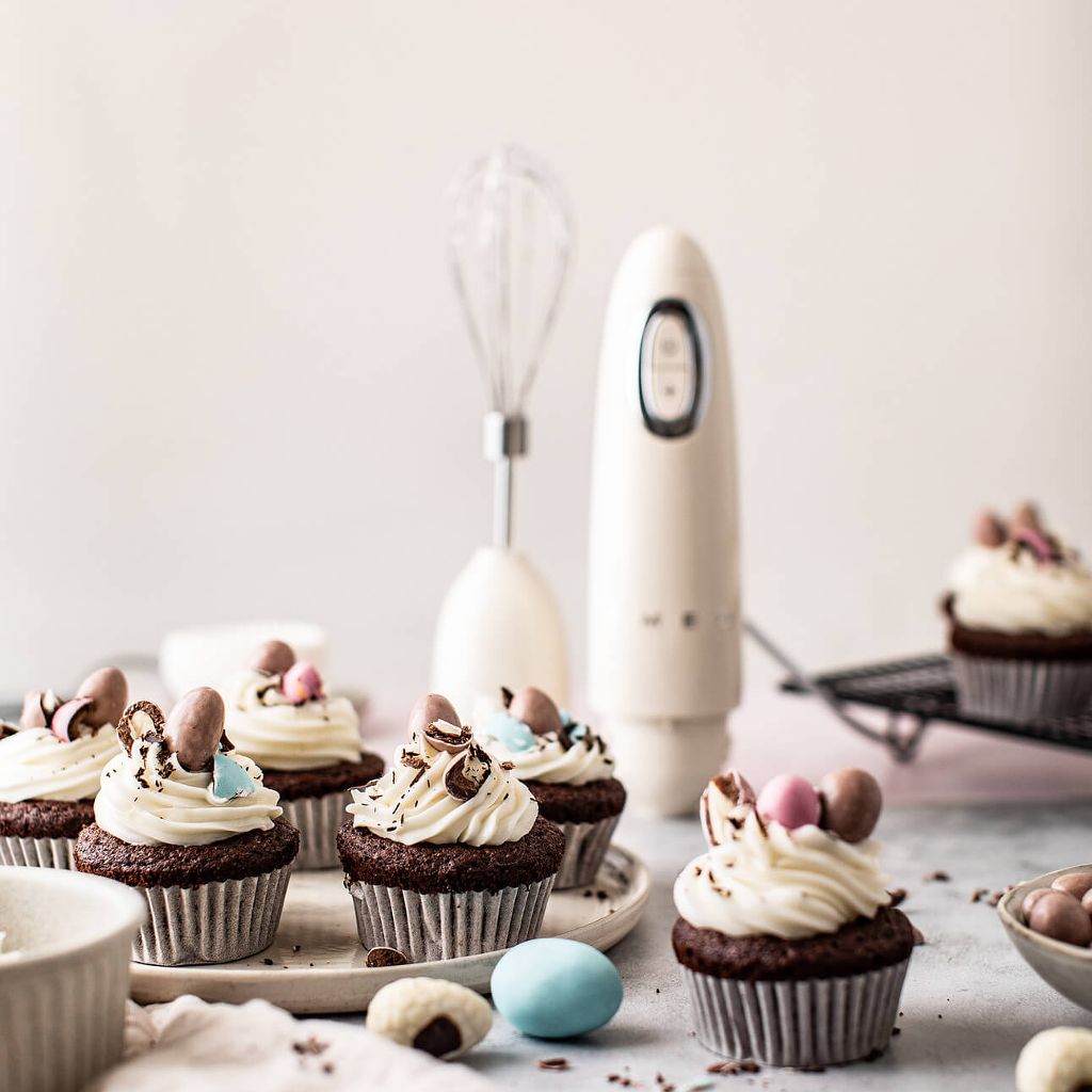 Chocolate cupcakes with vanilla frosting and candy eggs next to a cream-colored Smeg hand blender and whisk.