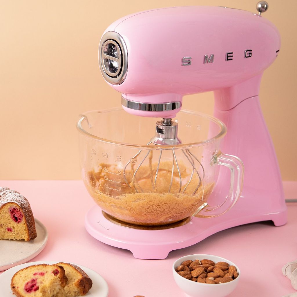 Pink Smeg stand mixer with a glass bowl next to a powdered raspberry bundt cake, almonds, and baking ingredients.