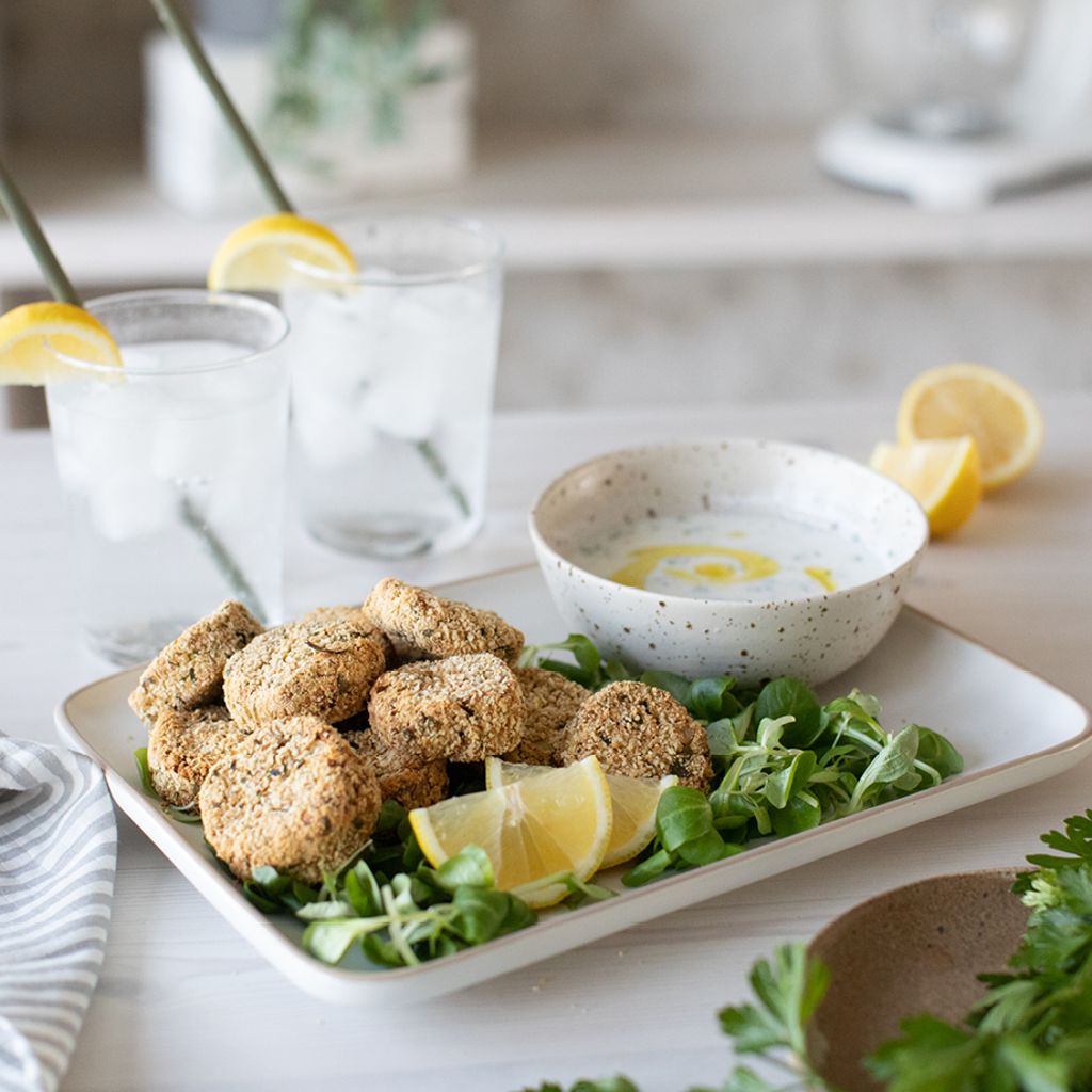 Crispy falafel patties on greens with a bowl of creamy dip, lemon wedges, and two iced drinks on a white table.