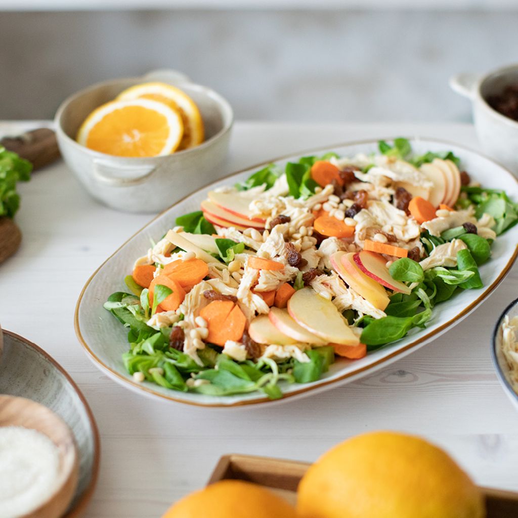 Platter of chicken salad with apple slices, carrots, and raisins, surrounded by bowls of fresh ingredients on a table.