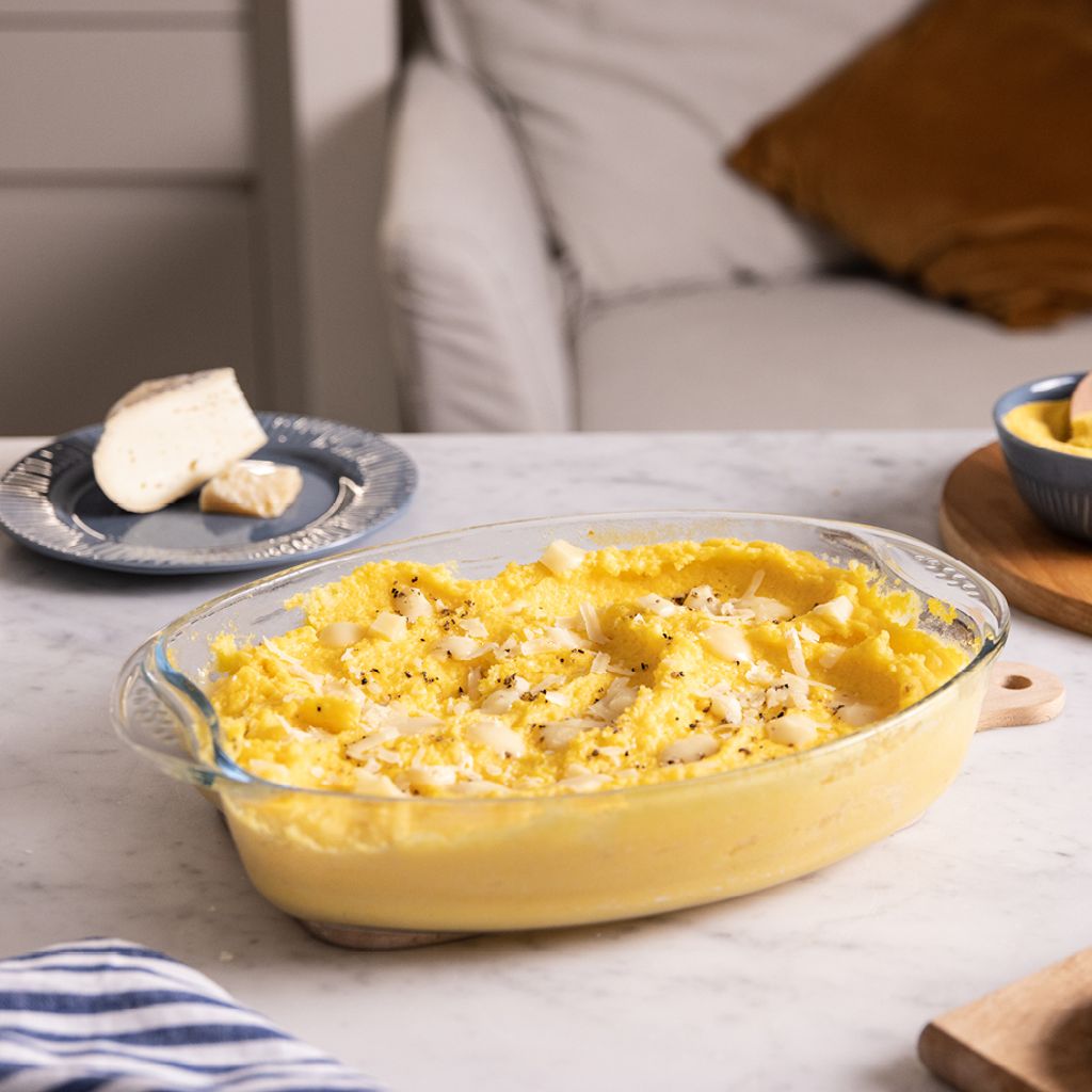 Creamy yellow polenta topped with melted cheese and pepper in a glass dish on a marble counter with ingredients.