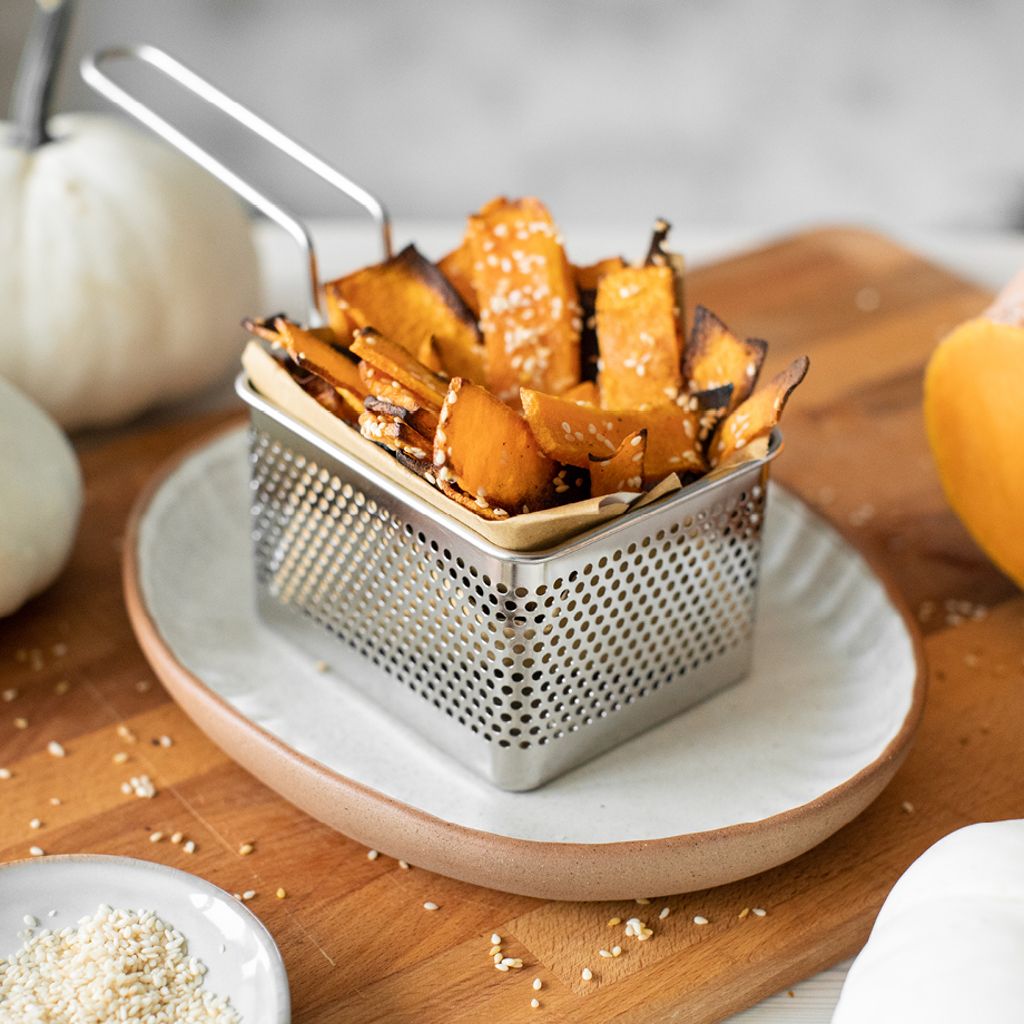 A small metal basket filled with thin, roasted pumpkin slices topped with sesame seeds on a wooden board.