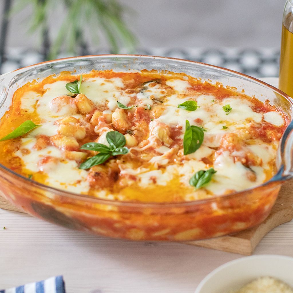 An oval glass baking dish filled with gnocchi in tomato sauce, topped with melted bubbling mozzarella cheese and fresh basil leaves.