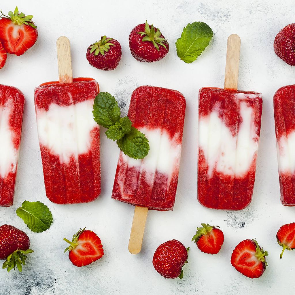 Five red and white swirled fruit popsicles arranged with fresh strawberries and mint leaves on a white surface.