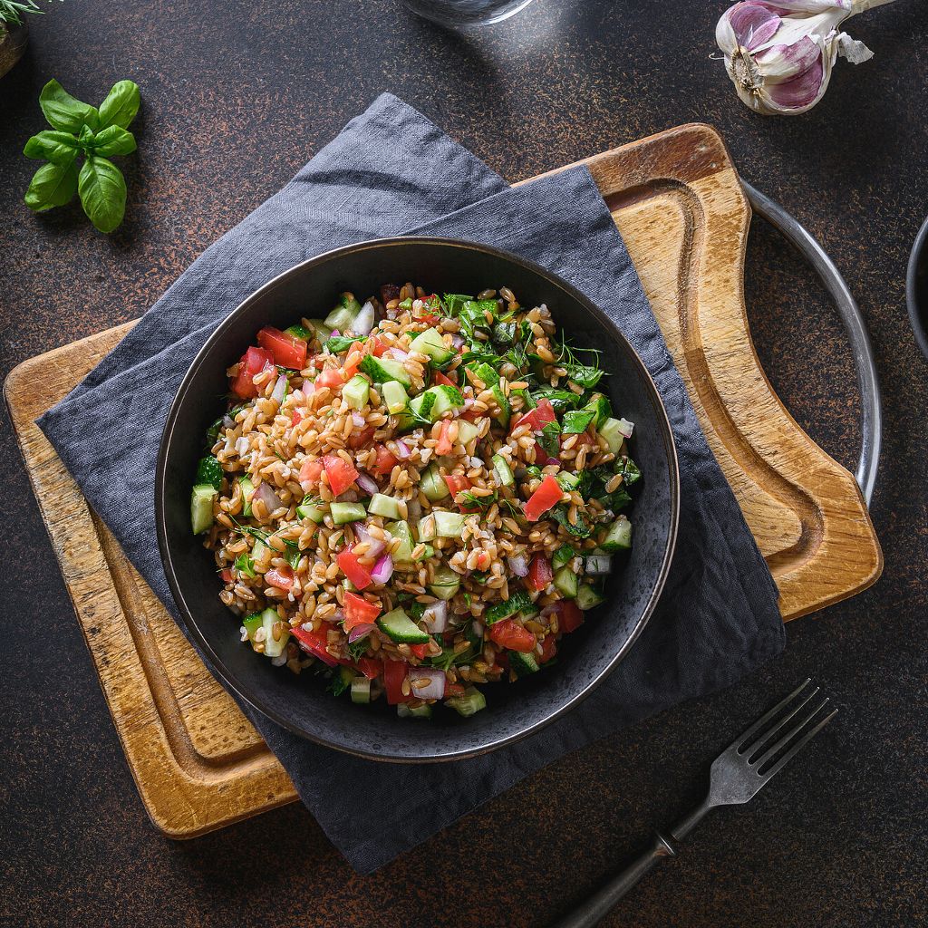 A dark bowl filled with cooked farro grains, diced tomatoes, cucumbers, red onion, and fresh green herbs.