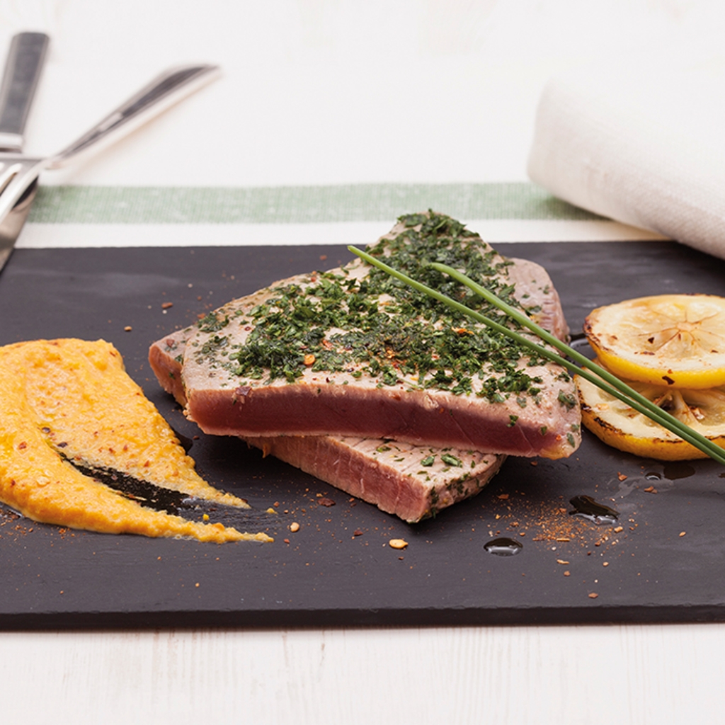 Seared tuna steaks topped with green herbs, served with a swipe of orange lentil cream and charred lemon on a black slate.