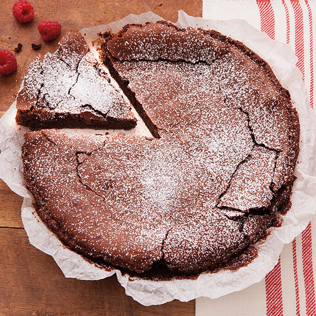 A sliced chocolate cake dusted with powdered sugar on parchment paper, topped with fresh raspberries on a wooden surface.
