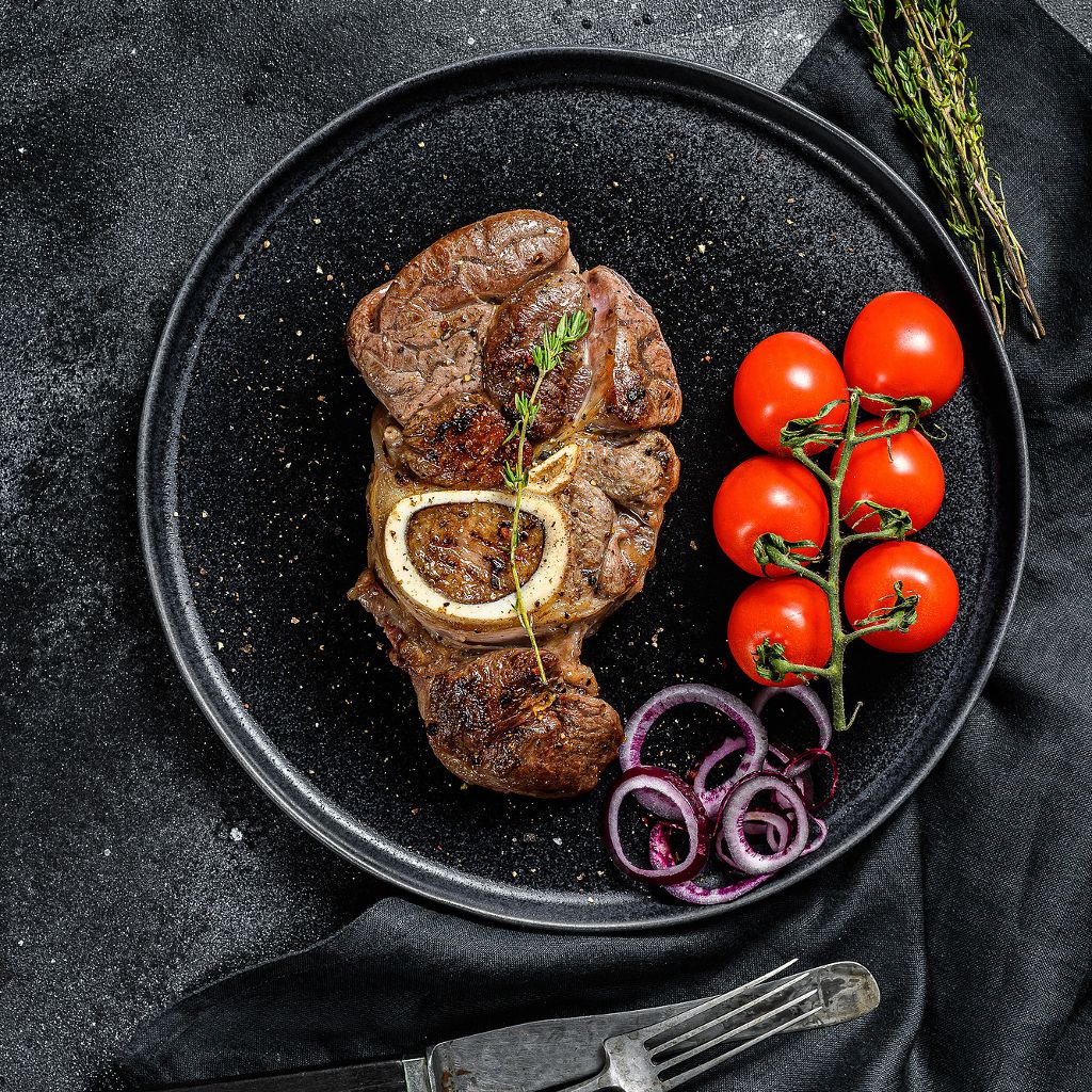 Braised veal shank served on a black plate with a vine of cherry tomatoes, red onion rings, and fresh herbs.