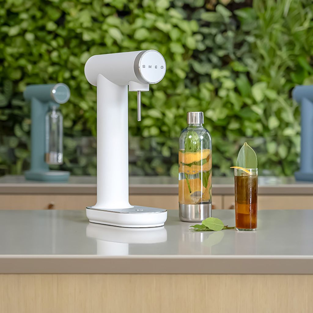 White Smeg soda maker with an infused water bottle and a dark-colored glass with garnish, on a white surface against a green background.