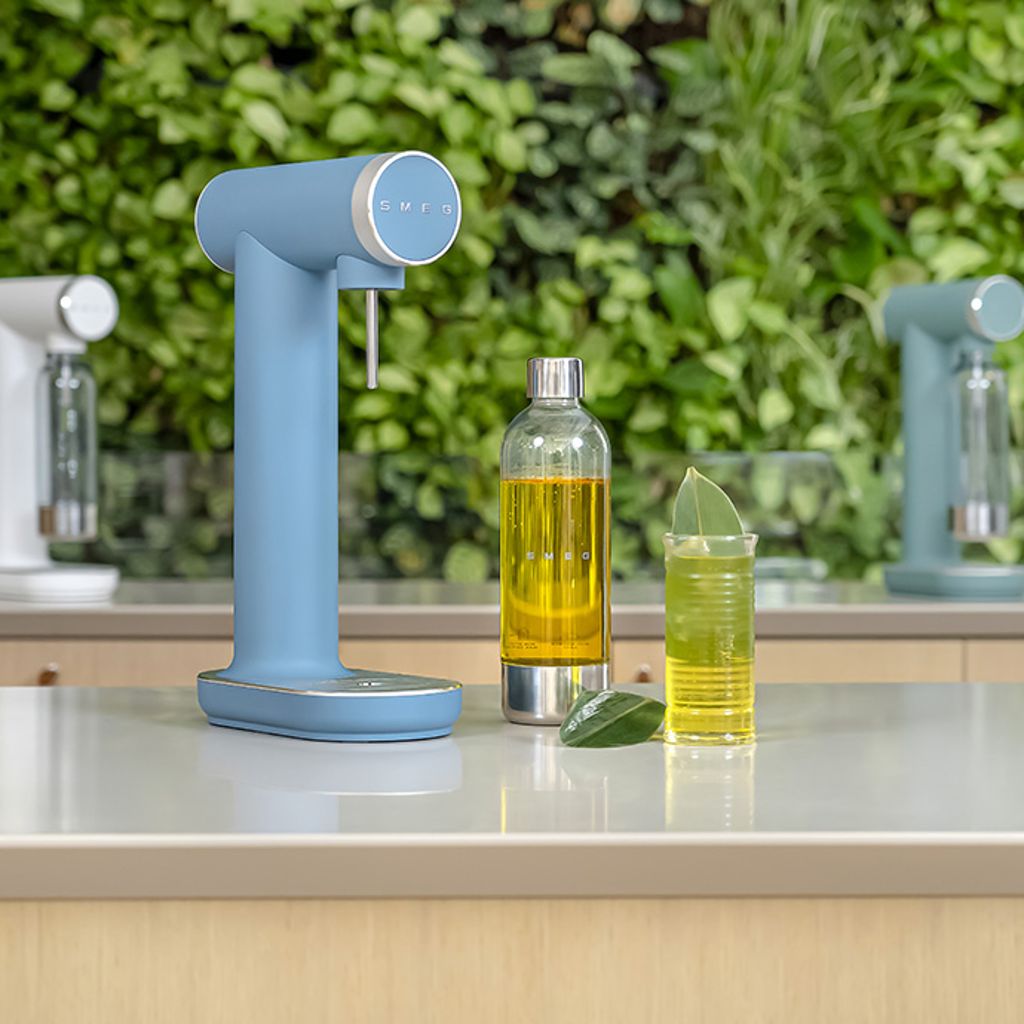 A blue Smeg soda maker with a bottle and a yellow glass on a white countertop against a backdrop of green plants.