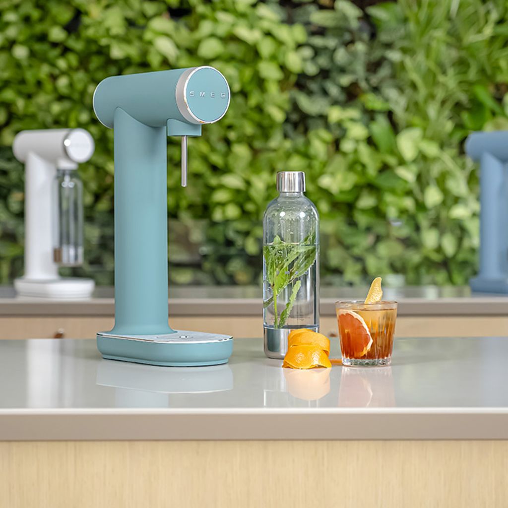 A matte Smeg soda maker with a bottle of water and mint, a glass with citrus fruit on a white surface, and a backdrop of green plants.