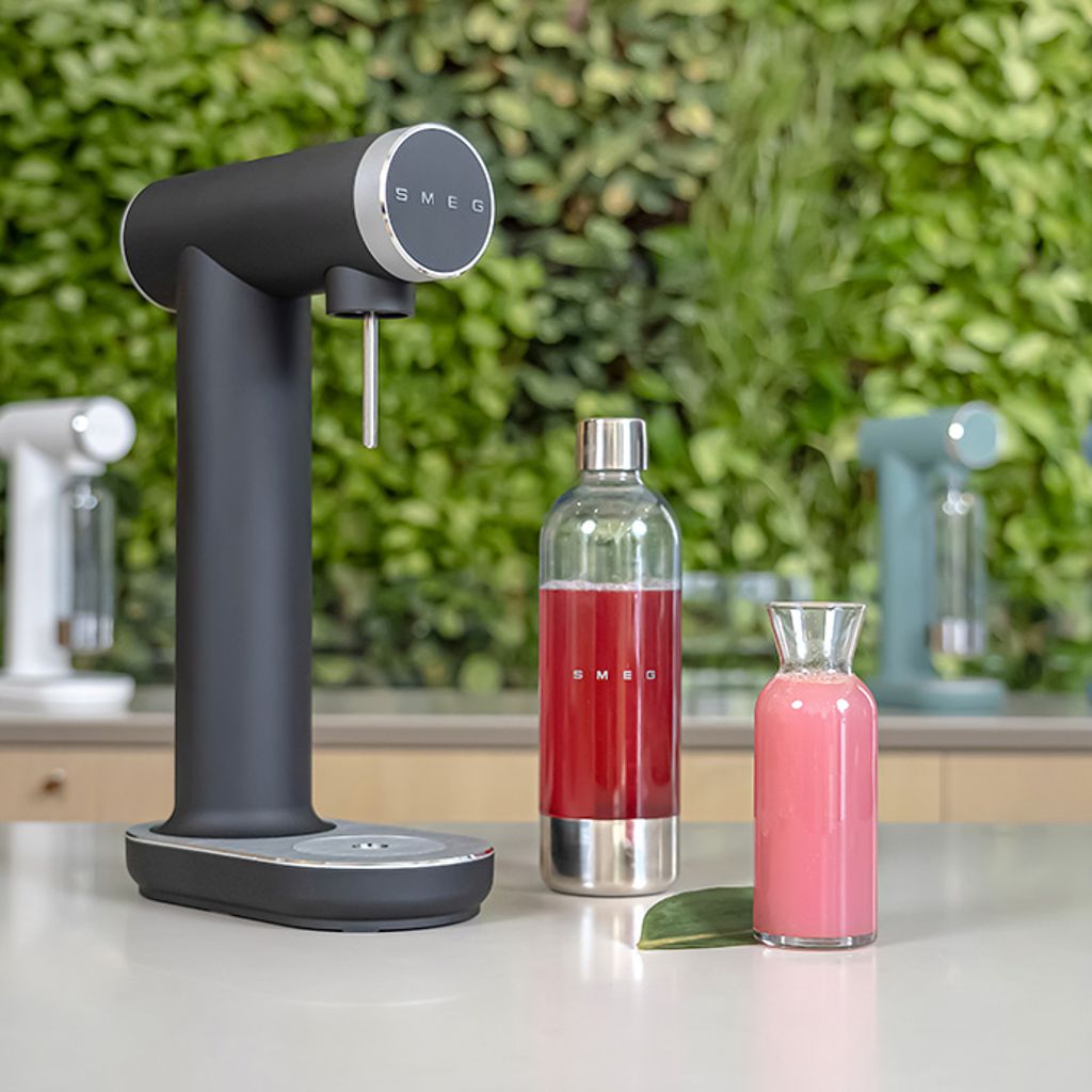 Smeg soda makers in various finishes, featuring a bottle and a carafe of pink drinks on a white surface against a green background.