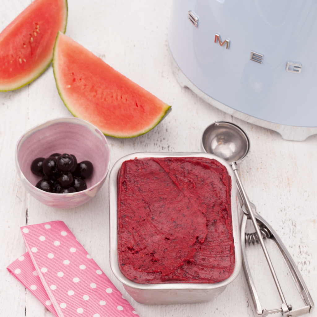 A container of dark red sorbet on a white table with an ice cream scoop, fresh cherries, and a watermelon slice.