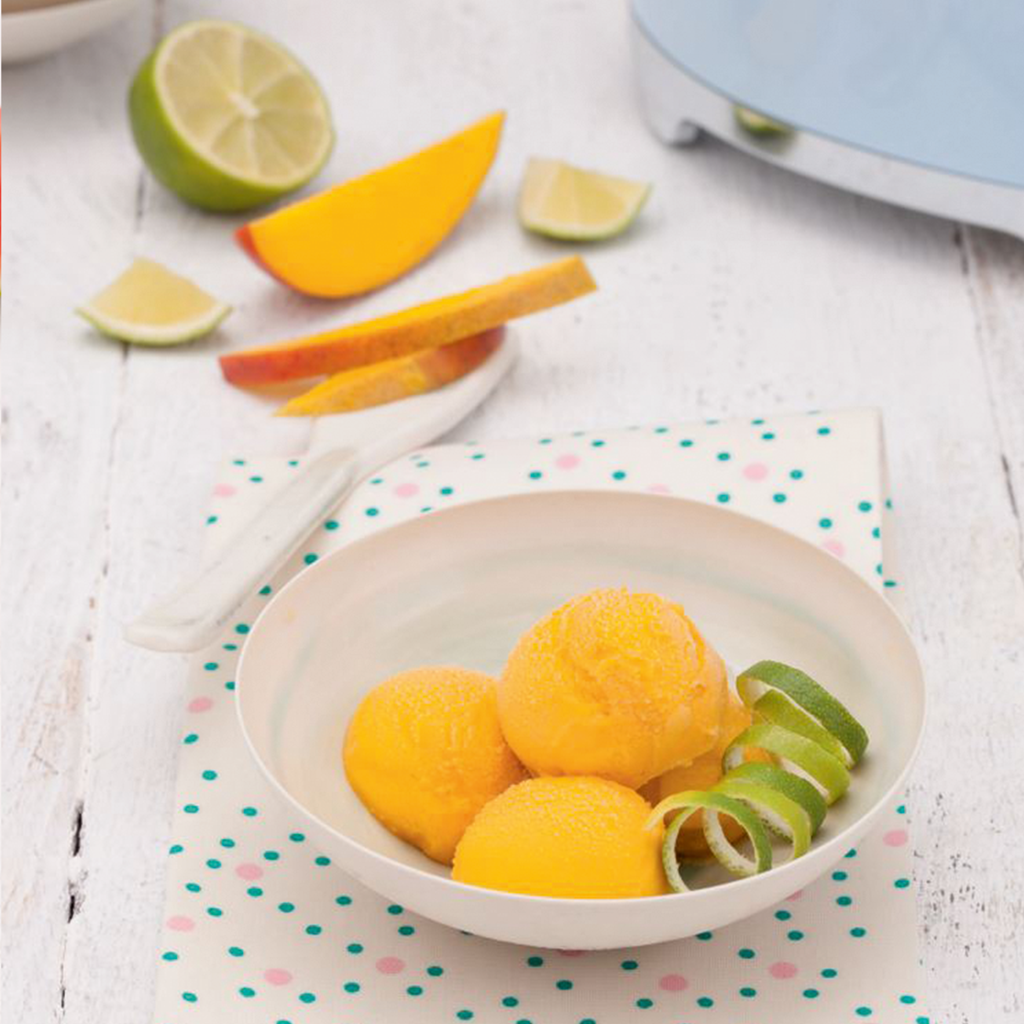 Three scoops of bright orange mango sorbet in a white bowl, garnished with a lime zest curl and fresh mango slices.