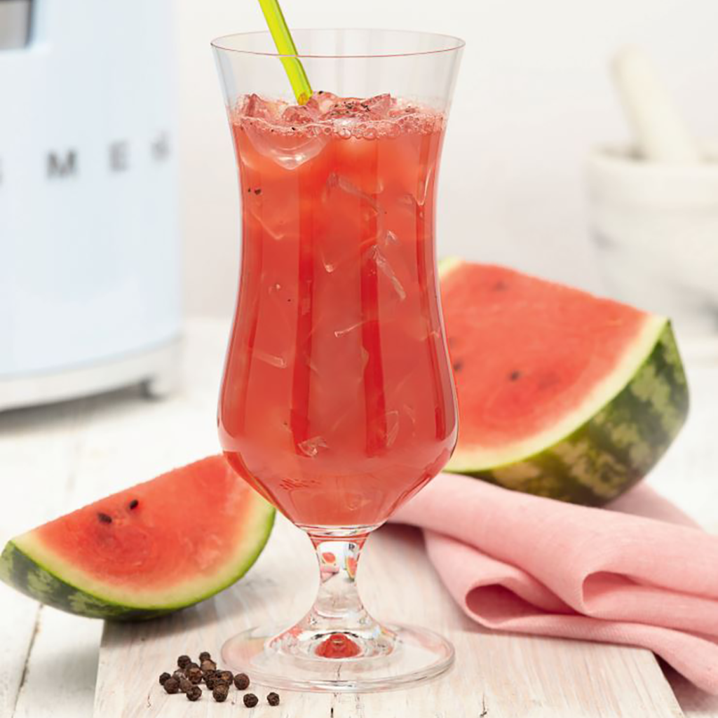 Three glasses of red watermelon juice garnished with black pepper and fresh watermelon wedges on a white table.