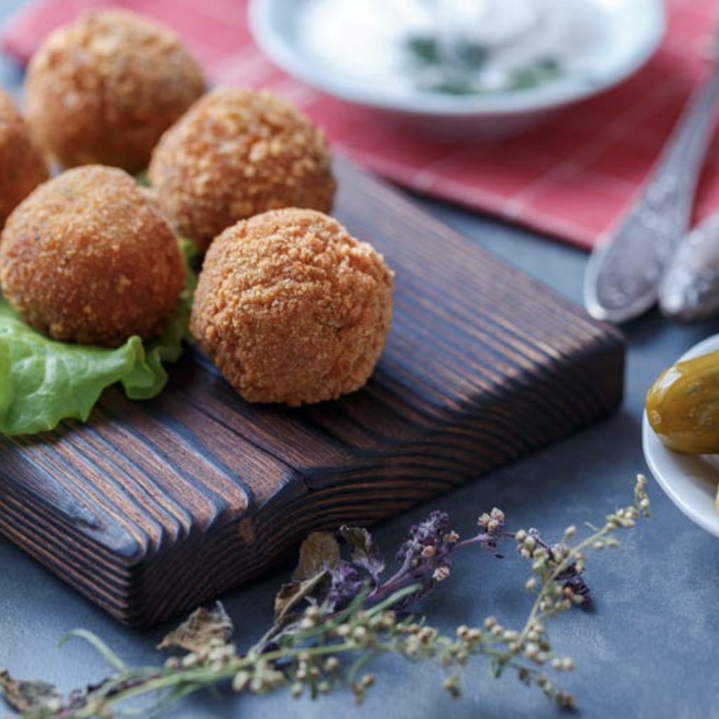 Six fried stuffed olives on a dark wooden board with lettuce, fresh herbs, and a small bowl of dipping sauce.